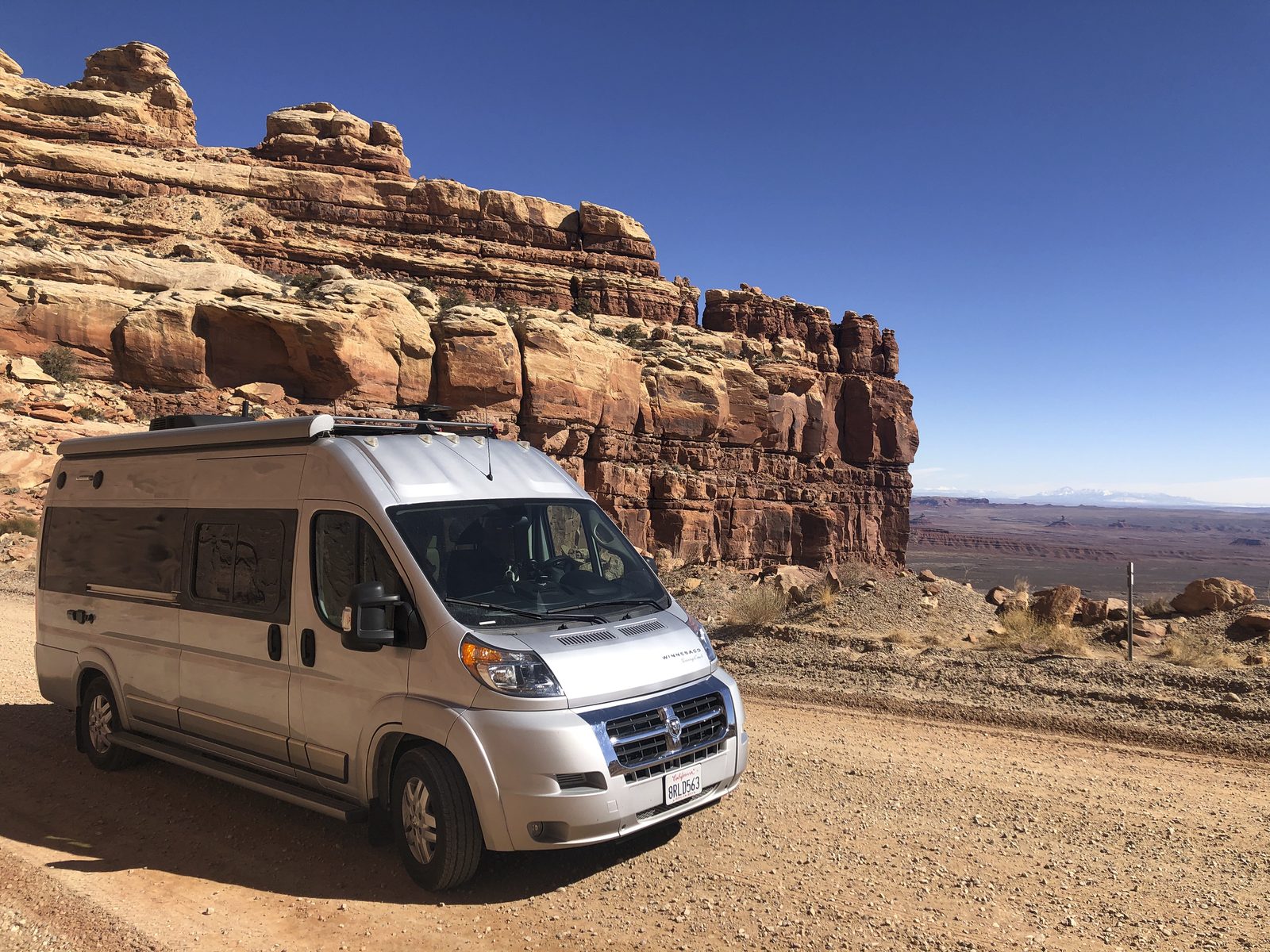 A silver high-roof campervan parked on a desert dirt road below red sandstone cliffs