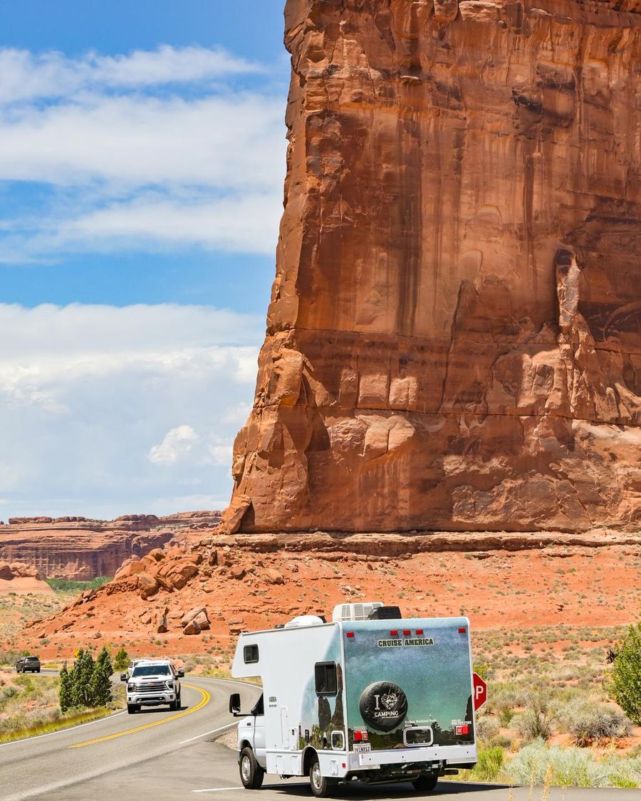 A rental Class C motorhome on a desert road below a tall red sandstone tower