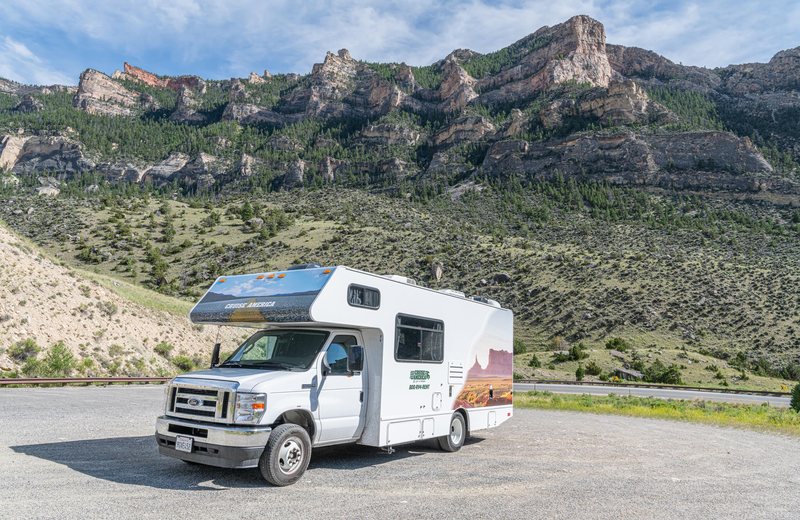 Class C rental motorhome parked at a gravel pullout below rocky, forested cliffs