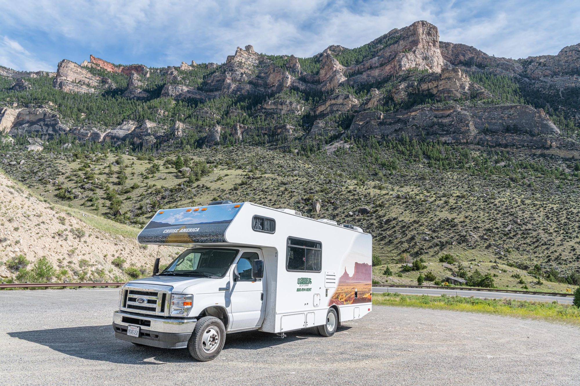 Class C rental motorhome parked at a gravel pullout below rocky, forested cliffs