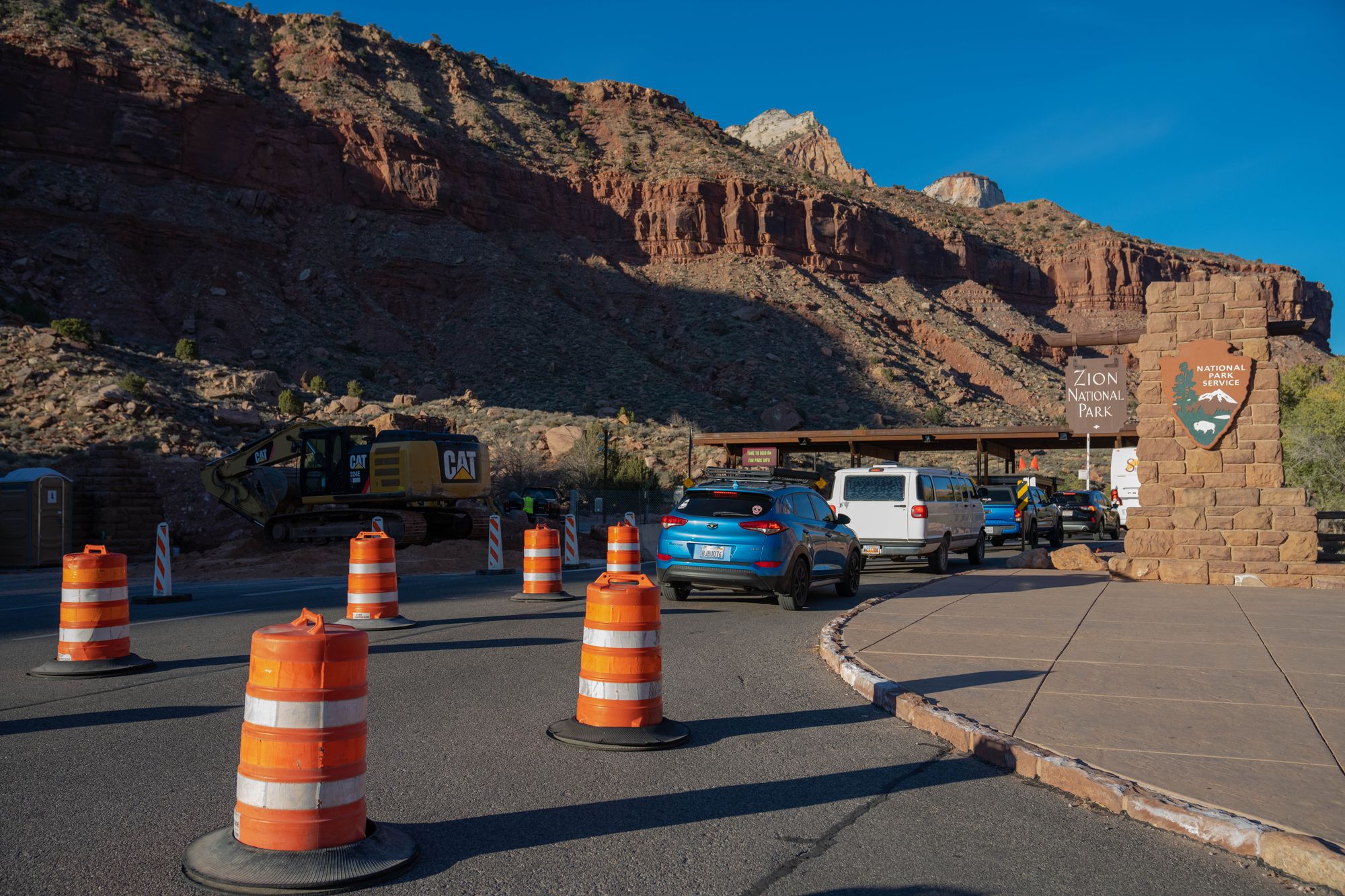 Cars queue between orange traffic barrels at the South Entrance of Zion National Park, with the stone park sign and red cliffs behind