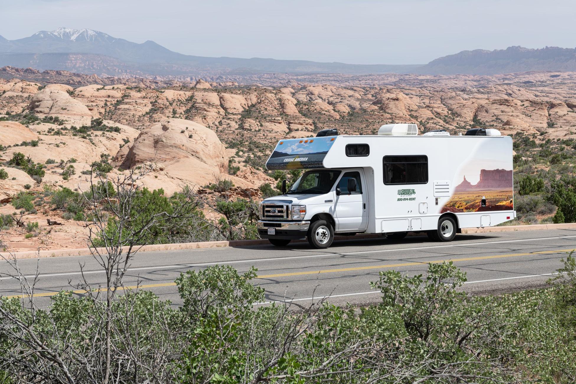 Class C motorhome on a desert road with slickrock domes and snowy mountains behind