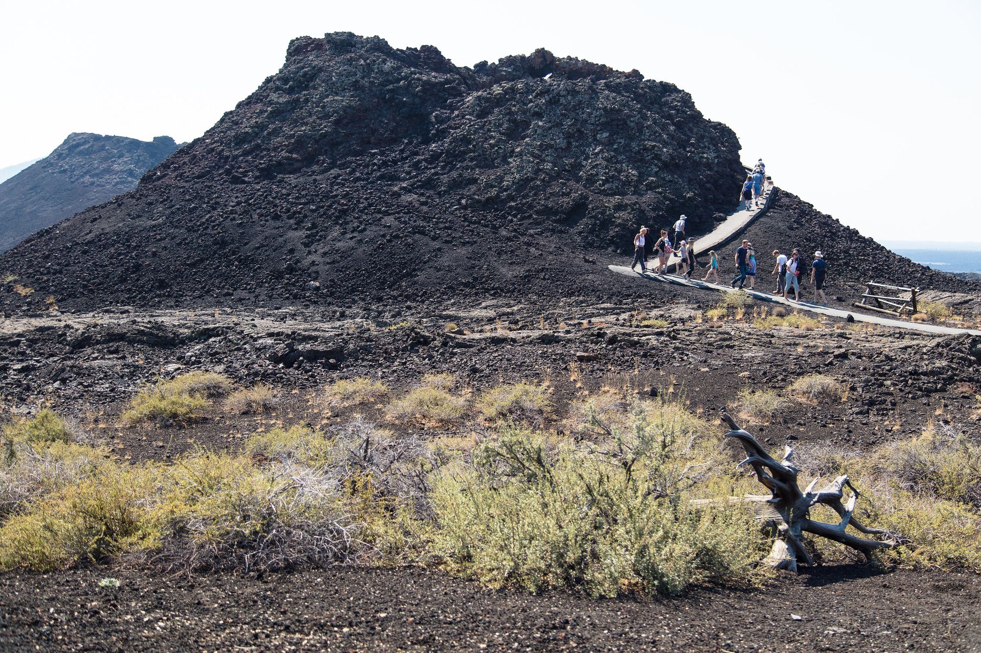 A paved trail leading across black lava past a steep, dark spatter cone at Craters of the Moon, with dry grass in front and visitors on the path