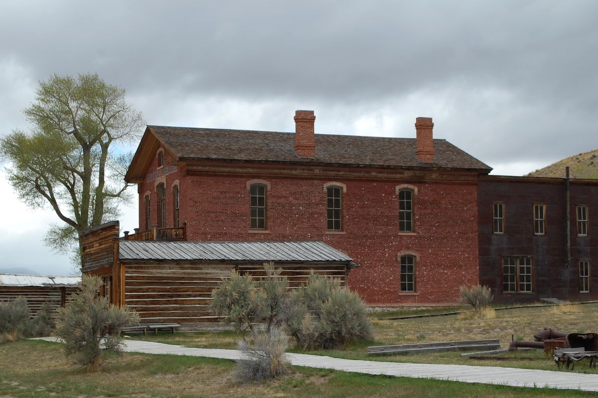 A large two-storey red brick building with a chimney and a small wooden porch, next to log buildings, in Bannack ghost town