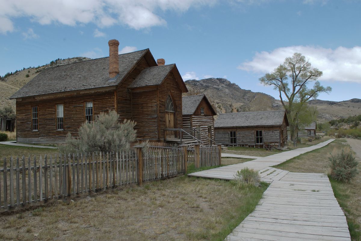 Weathered log and frame houses with steep roofs along a wooden boardwalk in Bannack, bare hills and blue sky behind