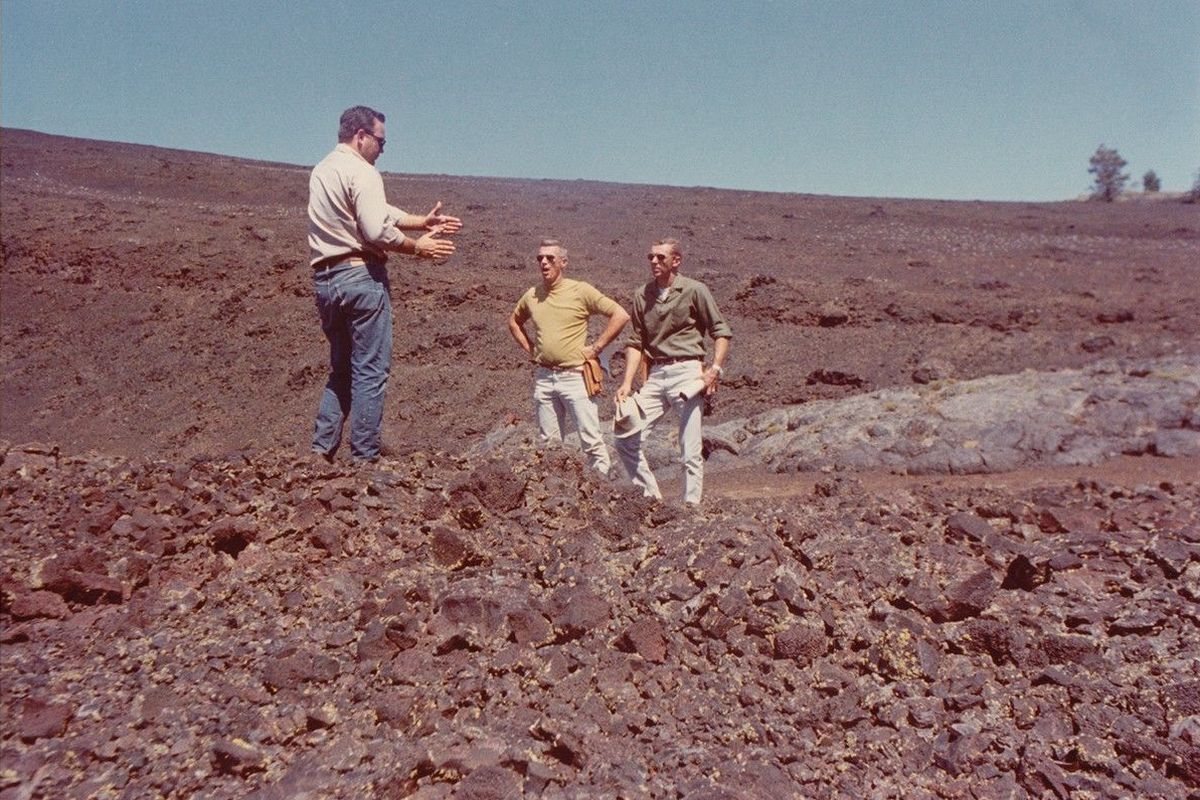 Three men in 1960s clothes stand on a rough, reddish lava field under a blue sky; one, gesturing, explains something to the other two