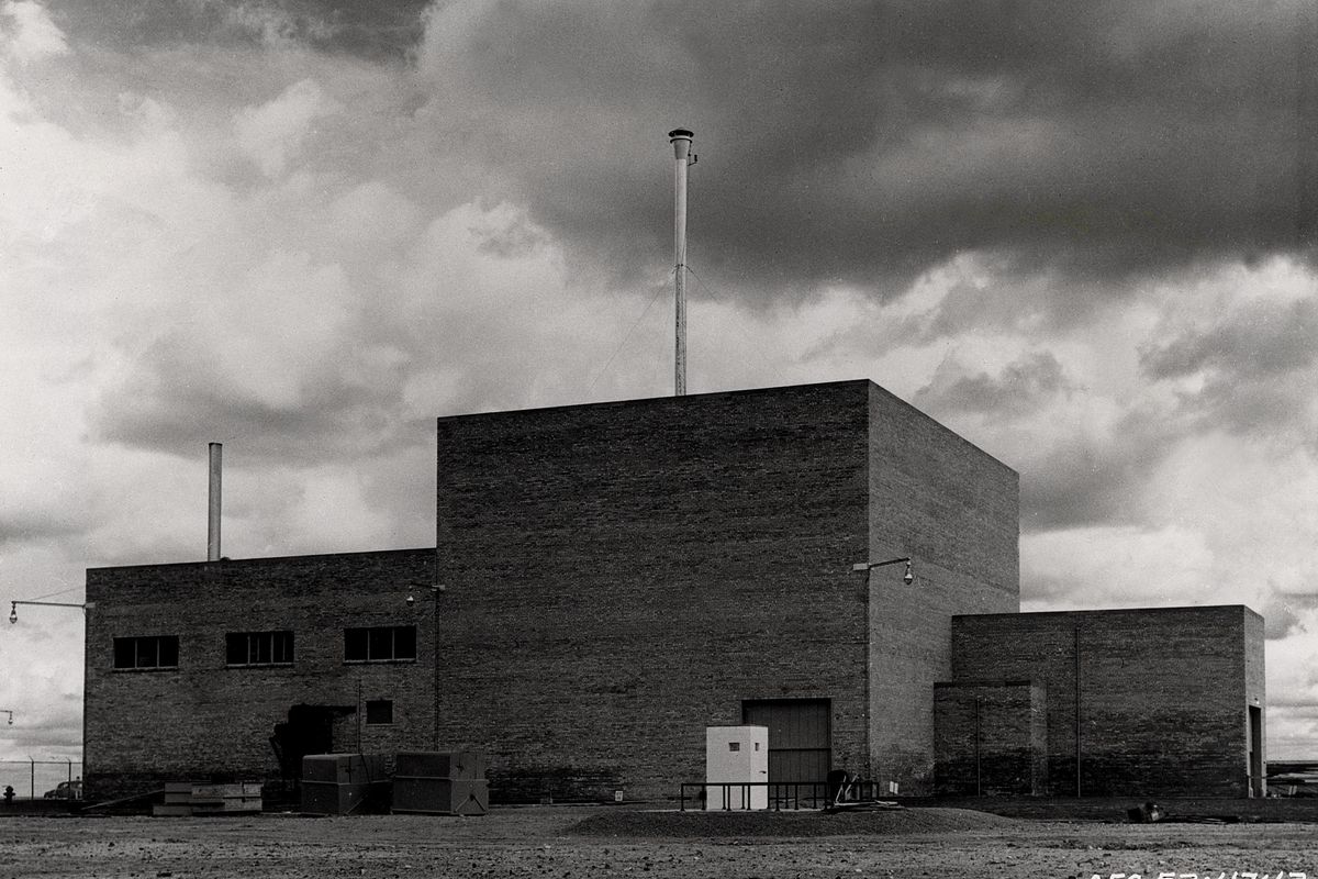 Black and white photo of the EBR-I building, a plain block-shaped reactor building with a tall stack, in flat desert under heavy clouds