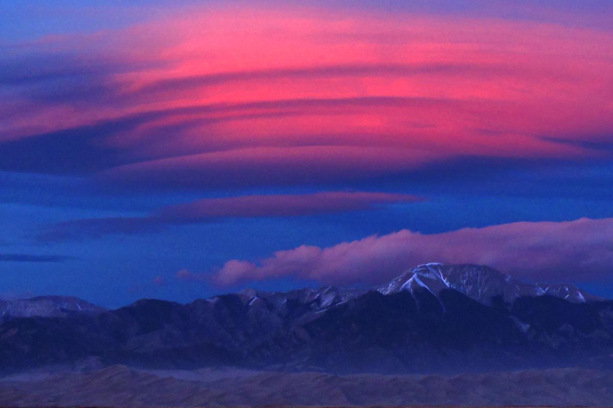 A large, pink, saucer-shaped lenticular cloud lit by the setting sun above the snow-capped Sangre de Cristo Mountains