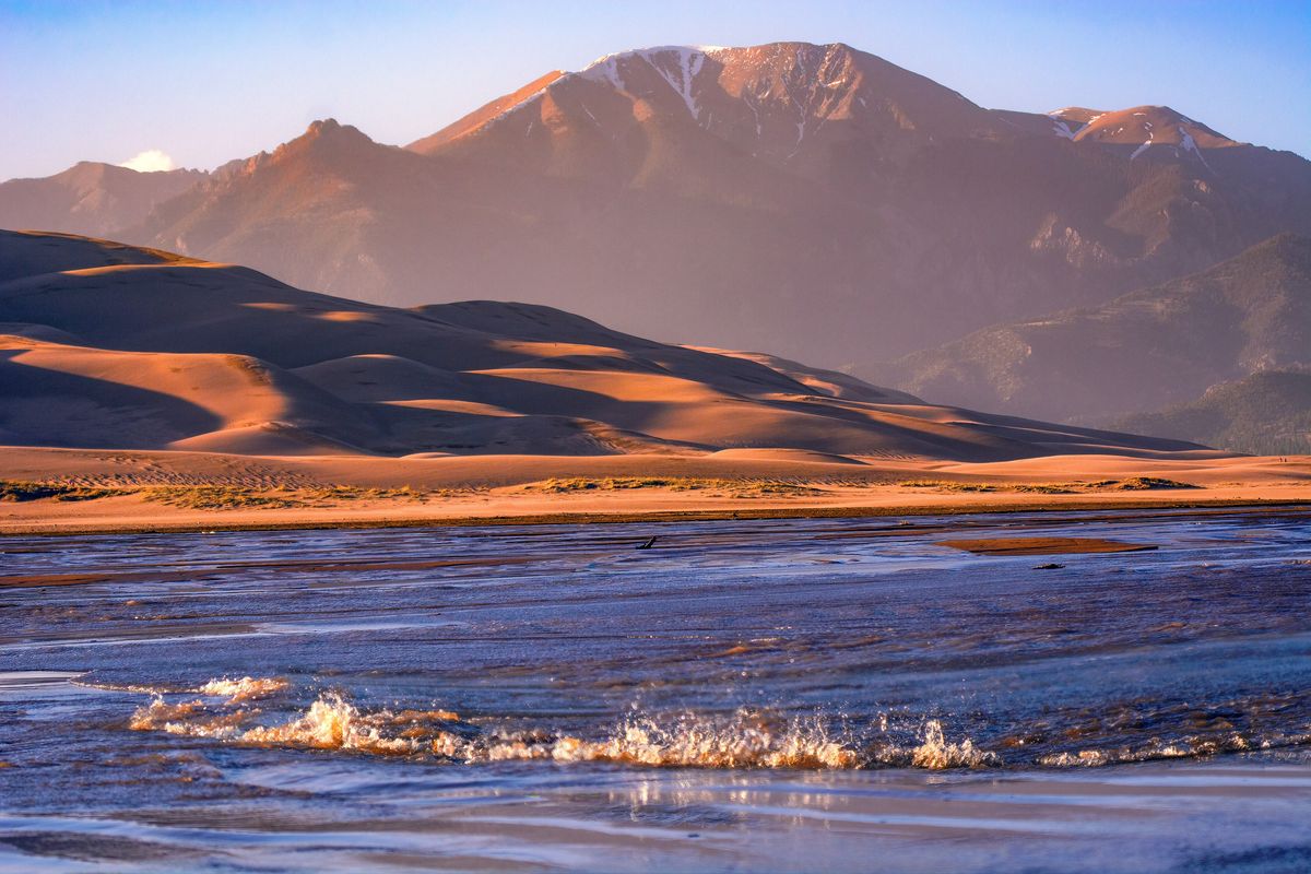 Medano Creek flowing in small waves across flat sand at sunrise, with a dune ridge and a mountain peak behind