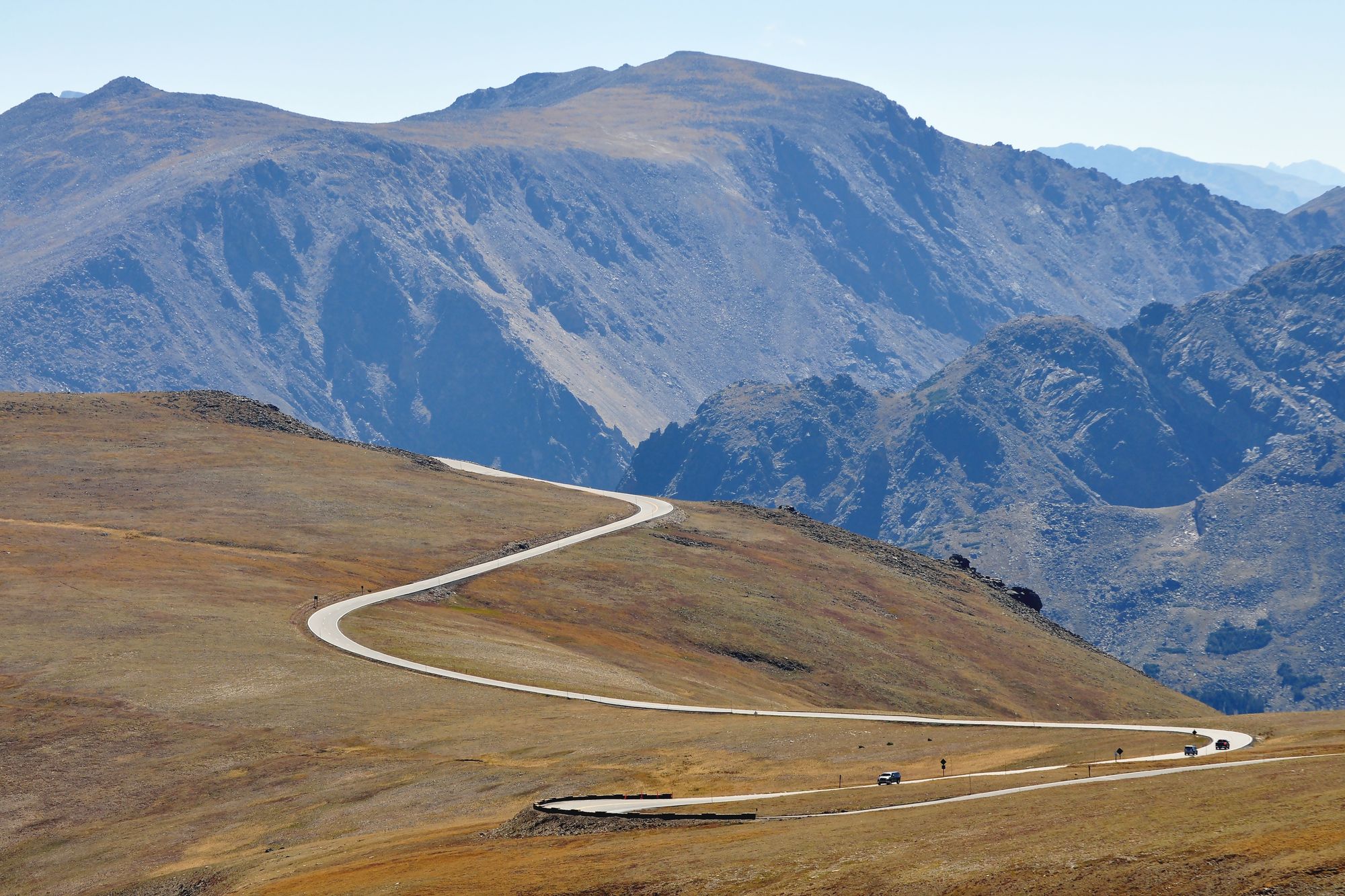 Trail Ridge Road curving across golden alpine tundra with a few cars on it, bare grey mountains behind, Rocky Mountain National Park