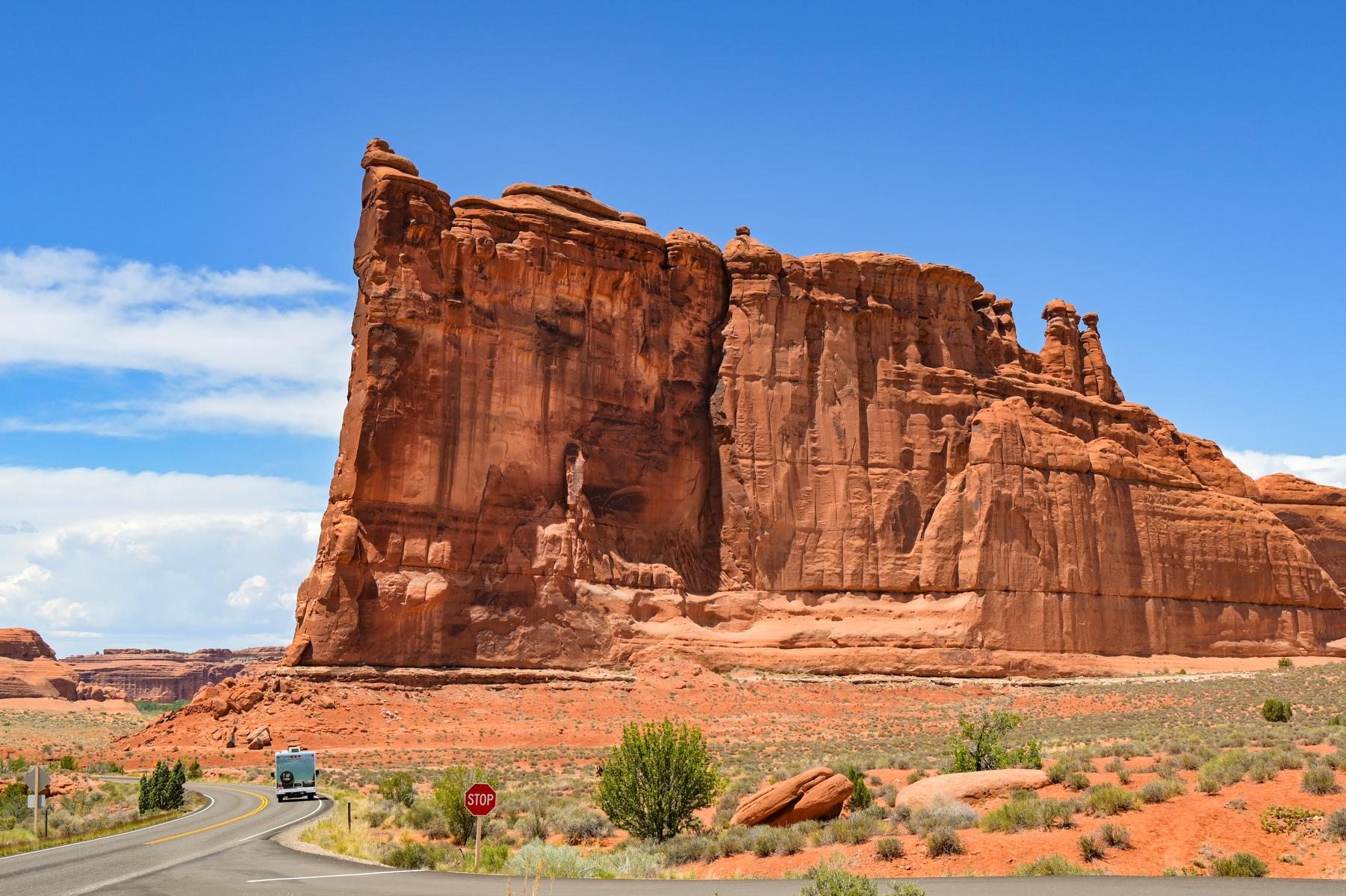 A road winding past towering red sandstone cliffs under a blue sky, a small RV on the road