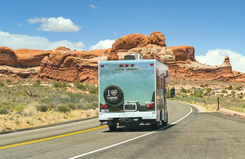 Class C motorhome driving a desert highway toward red sandstone formations