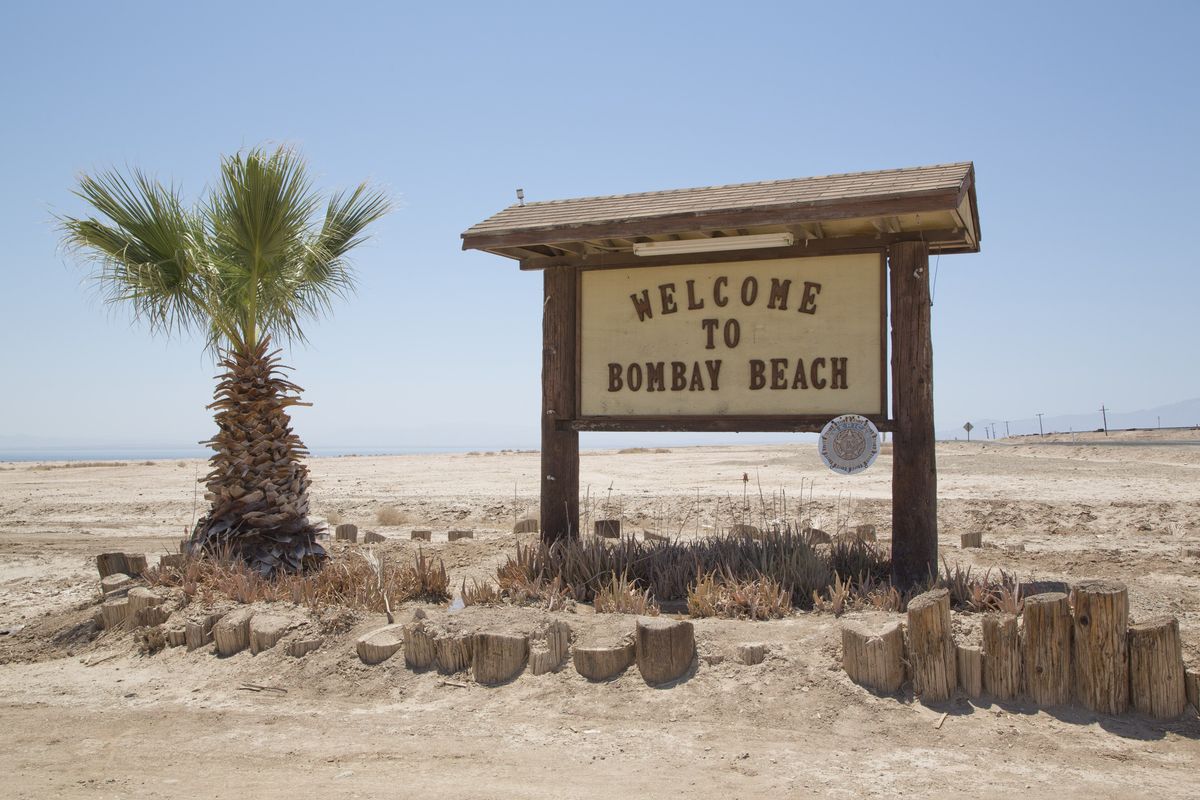 A wooden sign reading Welcome to Bombay Beach, with a palm tree beside it and flat, pale ground behind under a clear sky