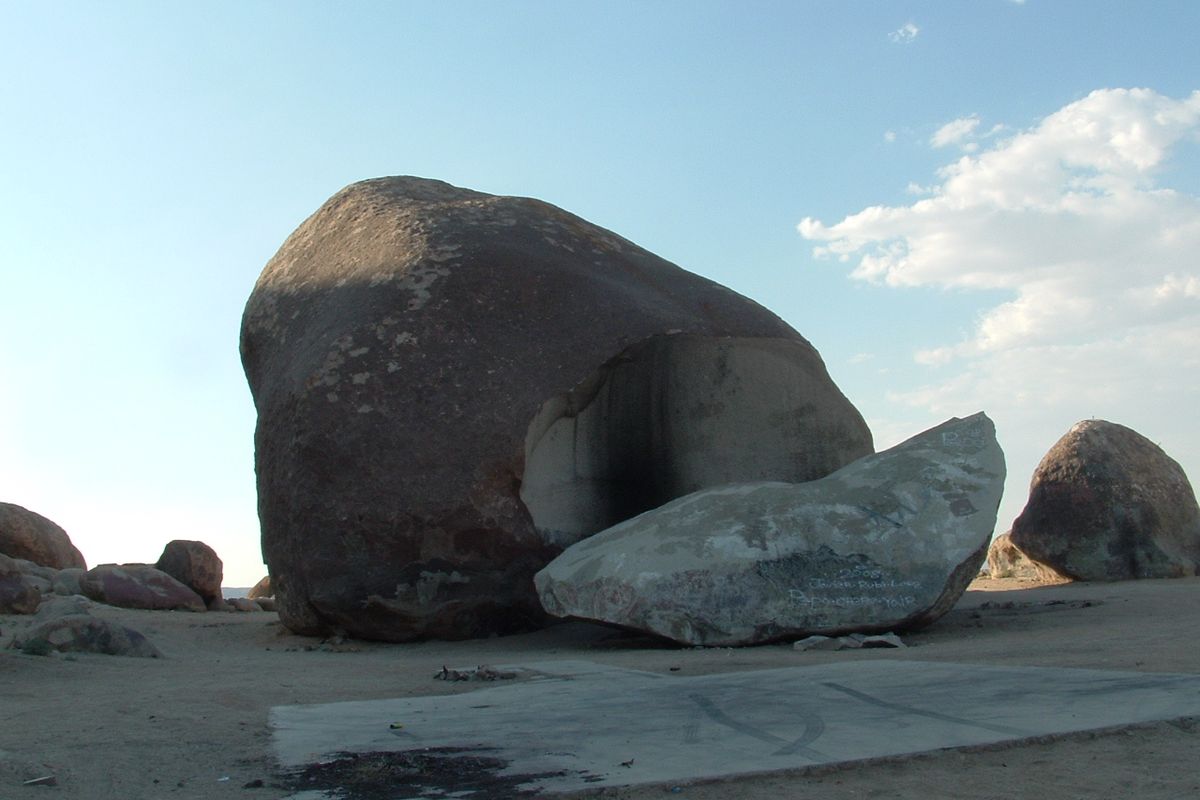 Giant Rock, a huge rounded granite boulder in the flat desert, with a large broken-off slab leaning in front of it and an old concrete pad in the foreground