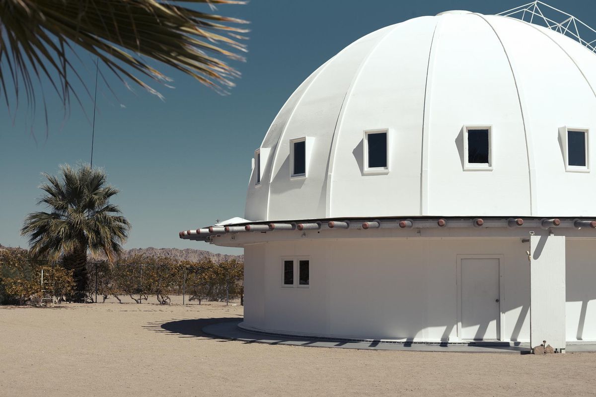 The white wooden dome of the Integratron, with a row of small square windows and a ring of white panels around its base, behind a gravel yard and a palm tree