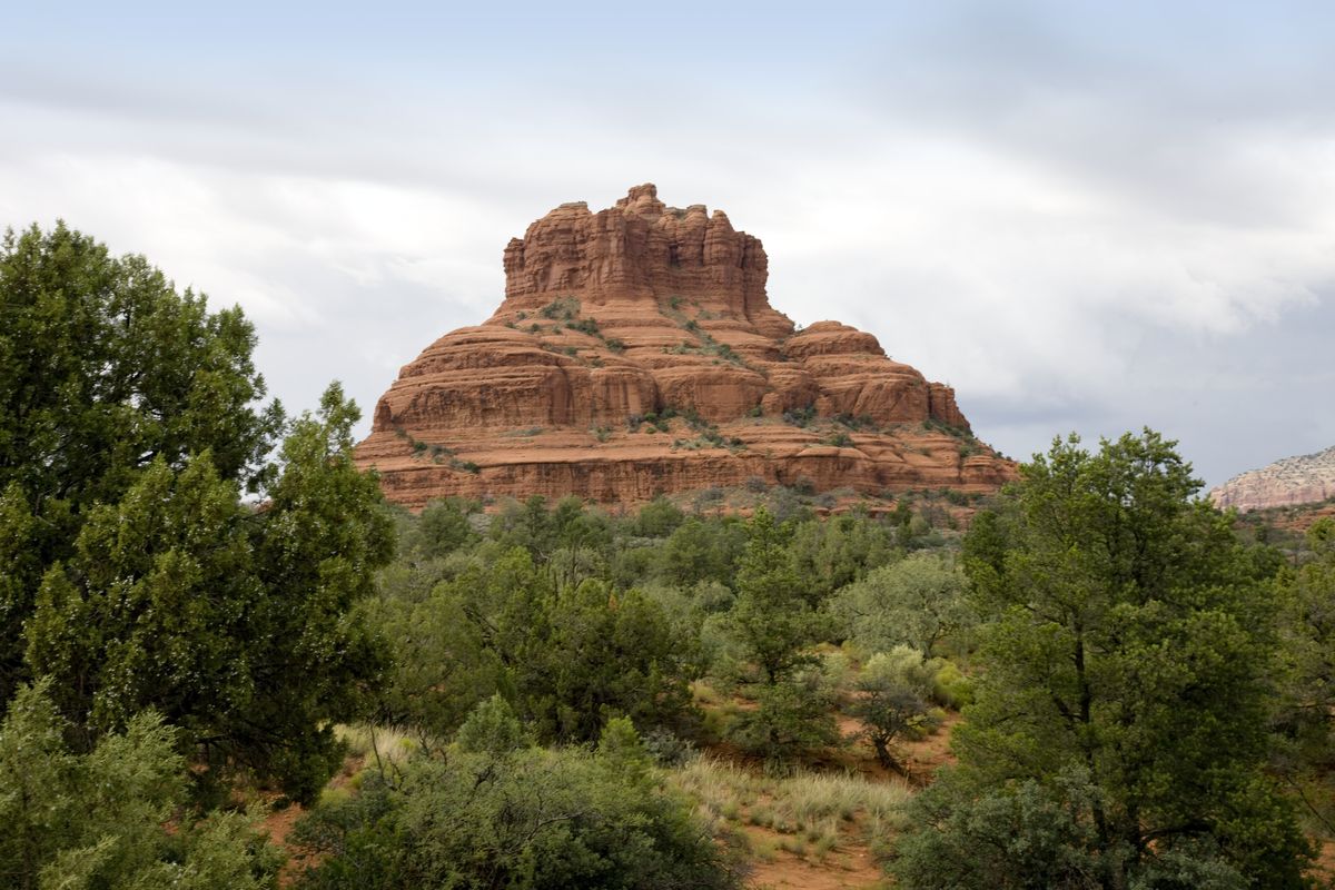 Bell Rock, a bell-shaped red sandstone butte near Sedona, Arizona, rising above green juniper and scrub under a cloudy sky