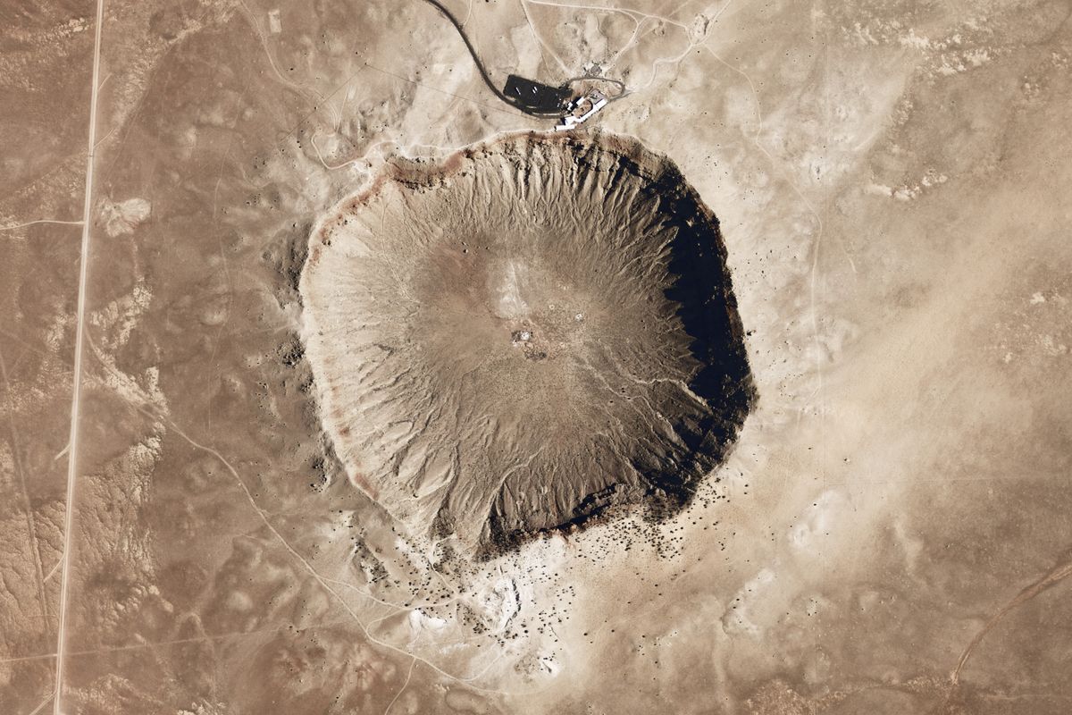 Satellite view of Meteor Crater from directly above: a round bowl with a pale rim and a road leading to the visitor centre on its north side