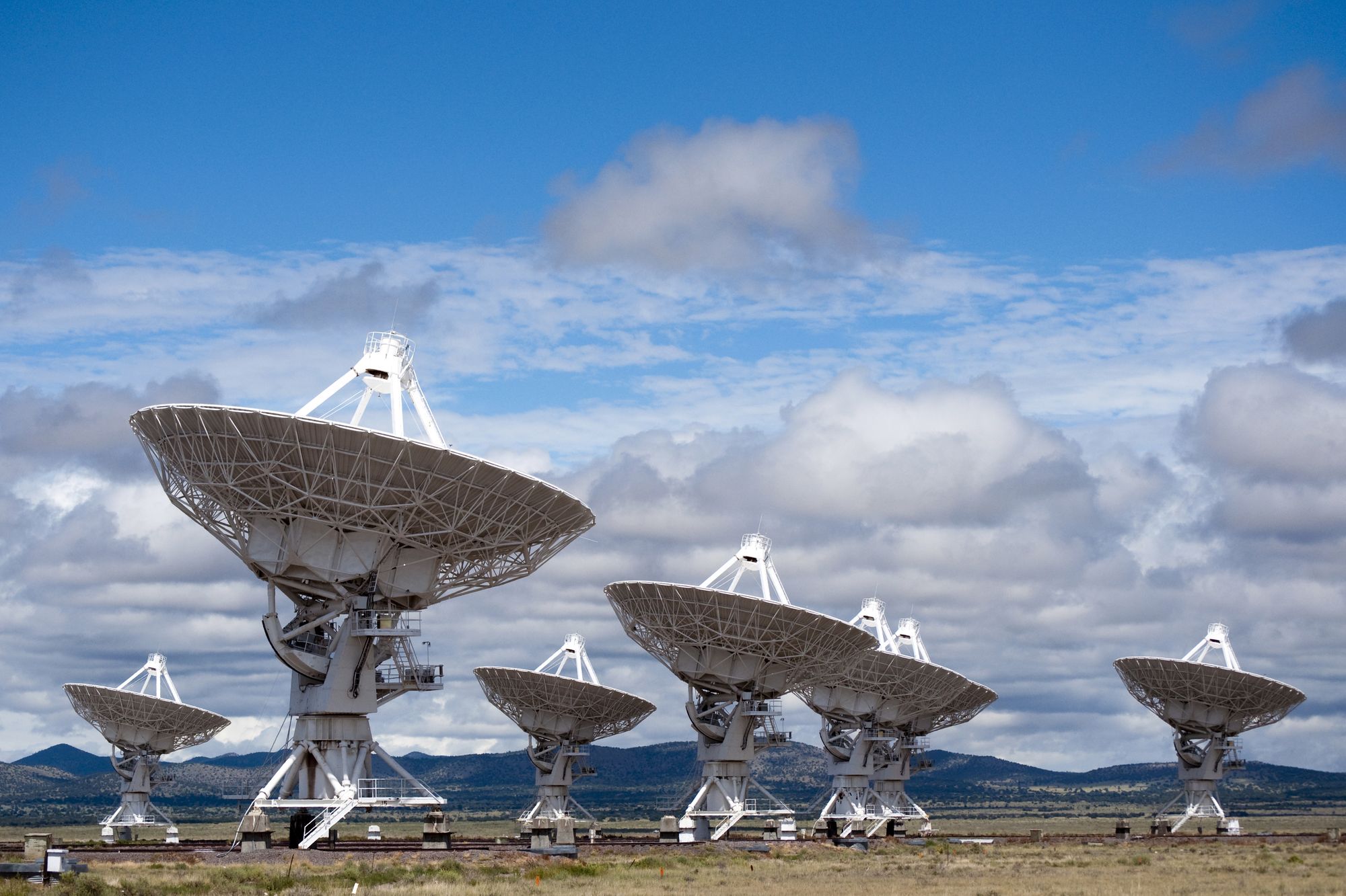 Several white radio telescope dishes of the Very Large Array on a plain in New Mexico, under a blue sky with white clouds