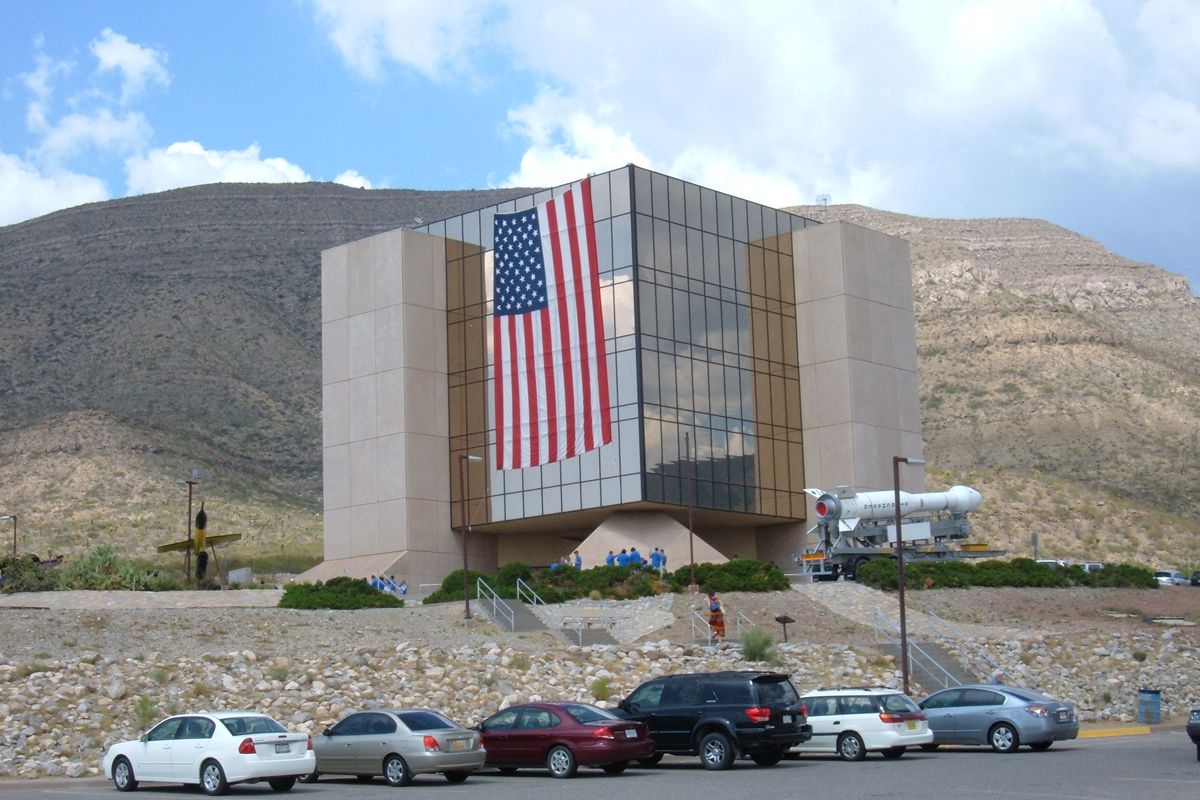 The tall glass-fronted building of the New Mexico Museum of Space History, with a large U.S. flag, on a hillside above Alamogordo
