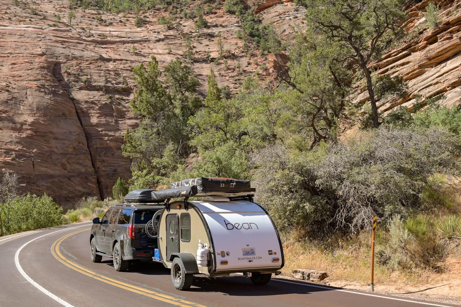 An SUV towing a small teardrop trailer on a curving road between red sandstone cliffs