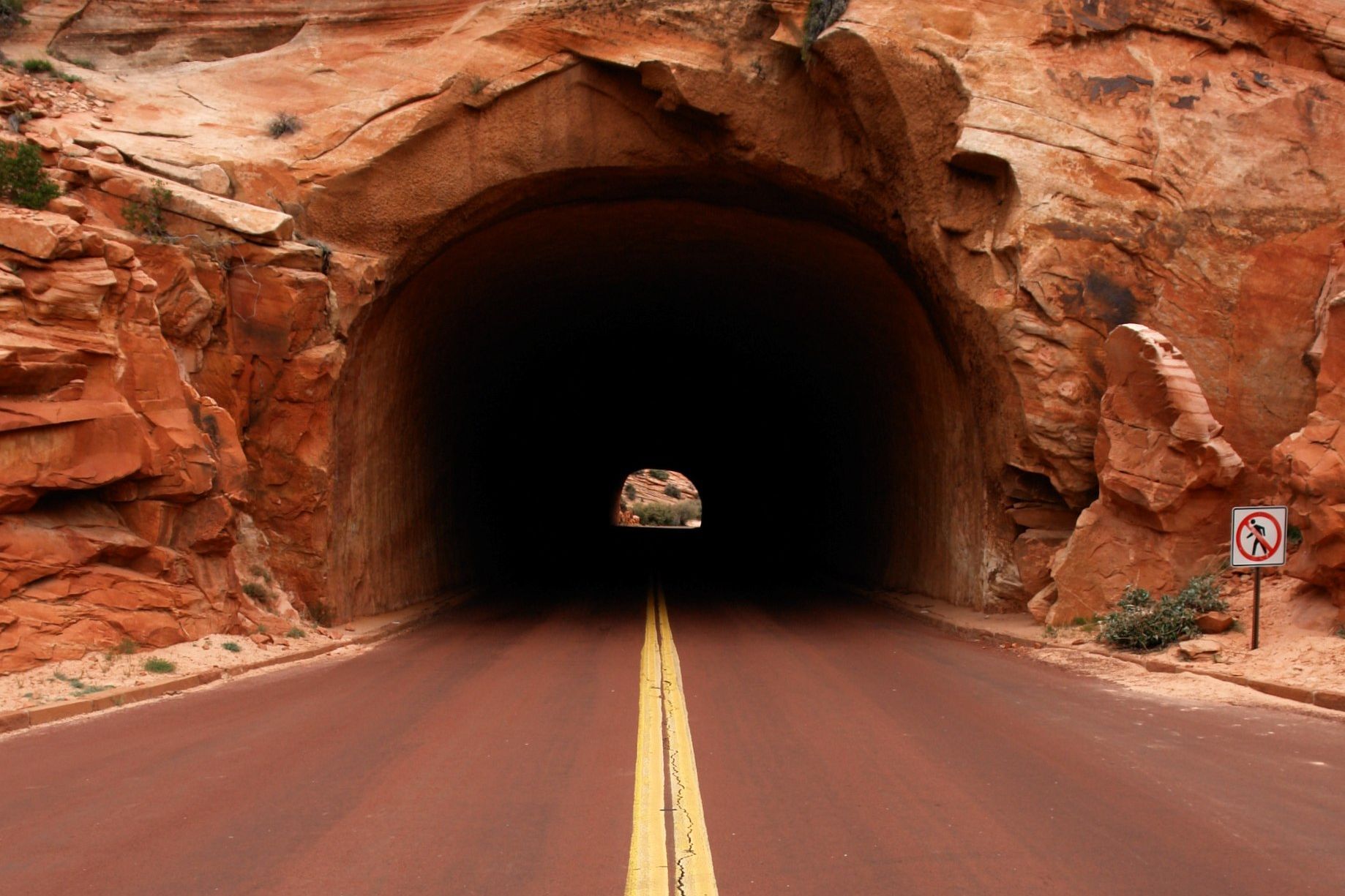 A two-lane road with a double yellow line running into a dark tunnel cut through orange sandstone on the Zion–Mount Carmel Highway
